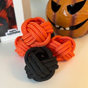 May include: Four decorative knot balls in a close-up shot. Three are vibrant orange, and one is black. The balls are made from a thick, woven rope. A carved pumpkin and a box with "Happy Halloween" text are in the background.