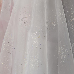 May include: White sheer fabric with scattered silver glitter. The fabric is draped, creating soft folds and shadows. The glitter catches the light, adding a sparkling effect. Ideal for bridal wear or decorative projects.