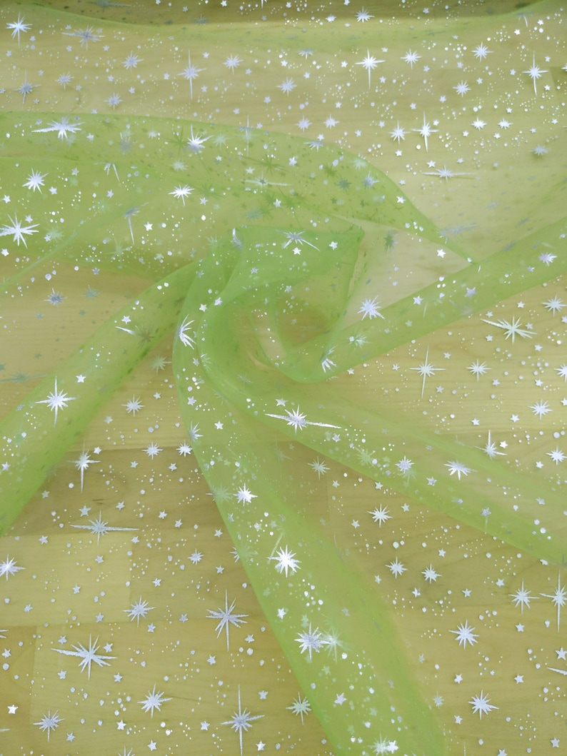 May include: Light green sheer fabric with a silver glitter star pattern. The fabric is draped and folded, showing the texture and sparkle of the material.