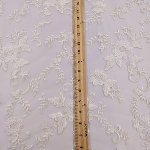 May include: Ivory lace fabric with floral embroidery. The fabric features a repeating pattern of flowers and leaves, with a scalloped edge. A wooden ruler shows the fabric is approximately 20 inches long.