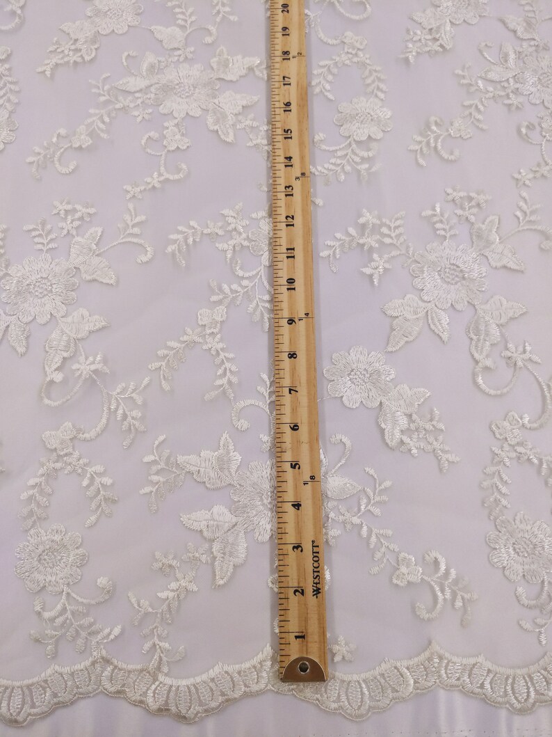 May include: Ivory lace fabric with floral embroidery. The fabric features a repeating pattern of flowers and leaves, with a scalloped edge. A wooden ruler shows the fabric is approximately 20 inches long.