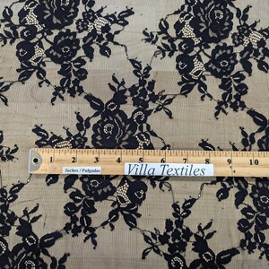 May include: Black floral lace fabric with a beige background. The fabric is measured with a ruler that reads "Villa Textiles" and "Inches/Pulgadas".