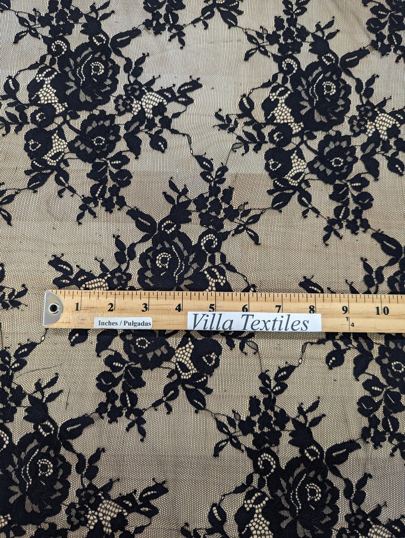 May include: Black floral lace fabric with a beige background. The fabric is measured with a ruler that reads "Villa Textiles" and "Inches/Pulgadas".