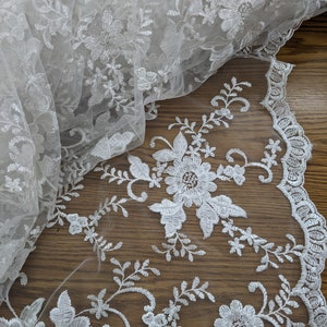May include: Ivory-colored floral lace fabric. The delicate lace features embroidered flowers and leaves, with a scalloped edge. The fabric is partially rolled, showcasing the intricate details and texture. Ideal for bridal wear or decorative projects.