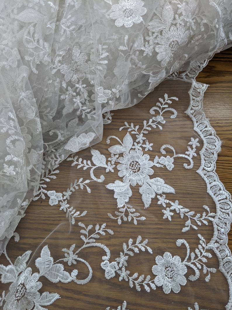 May include: Ivory-colored floral lace fabric. The delicate lace features embroidered flowers and leaves, with a scalloped edge. The fabric is partially rolled, showcasing the intricate details and texture. Ideal for bridal wear or decorative projects.