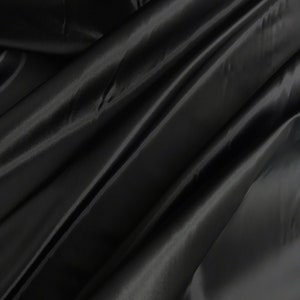 May include: Black satin fabric with a smooth, silky texture. The fabric is draped and folded, creating soft, flowing lines.