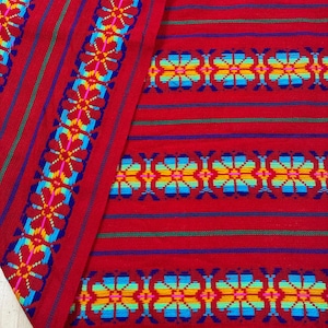 May include: A vibrant red textile featuring horizontal stripes and repeating floral patterns in bright shades of blue, yellow, green, and pink. The fabric displays a dense, woven texture with intricate geometric flower motifs.