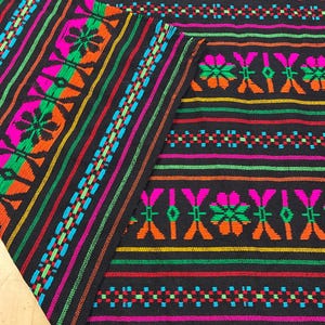 May include: A black textile featuring horizontal stripes and intricate geometric patterns in vibrant neon pink, bright orange, electric green, and cyan blue. The fabric displays repeating floral-like motifs and small diamond shapes.