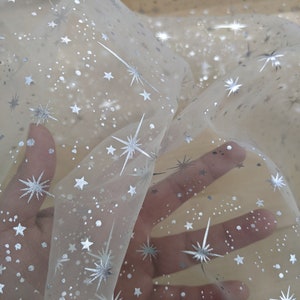 May include: White sheer fabric with a silver star pattern. The fabric is translucent and has a delicate, sparkly look.