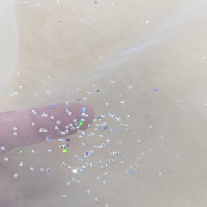 May include: Close-up of a sheer, cream-coloured fabric with scattered iridescent glitter. The glitter particles are small and reflect light, creating a sparkling effect. The fabric appears delicate and airy, with a soft, translucent quality.