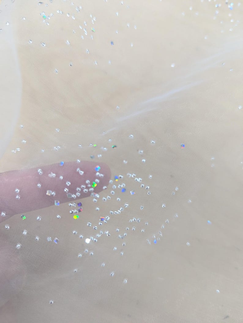 May include: Close-up of a sheer, cream-coloured fabric with scattered iridescent glitter. The glitter particles are small and reflect light, creating a sparkling effect. The fabric appears delicate and airy, with a soft, translucent quality.