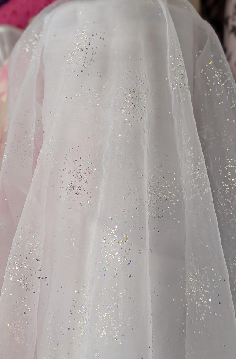 May include: White sheer fabric with scattered silver glitter. The fabric is draped, creating soft folds and shadows. The glitter catches the light, adding a sparkling effect. Ideal for bridal wear or decorative projects.
