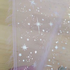 May include: A sheer white fabric with a silver glitter pattern of stars and dots. The fabric is folded in half and the edge is frayed.