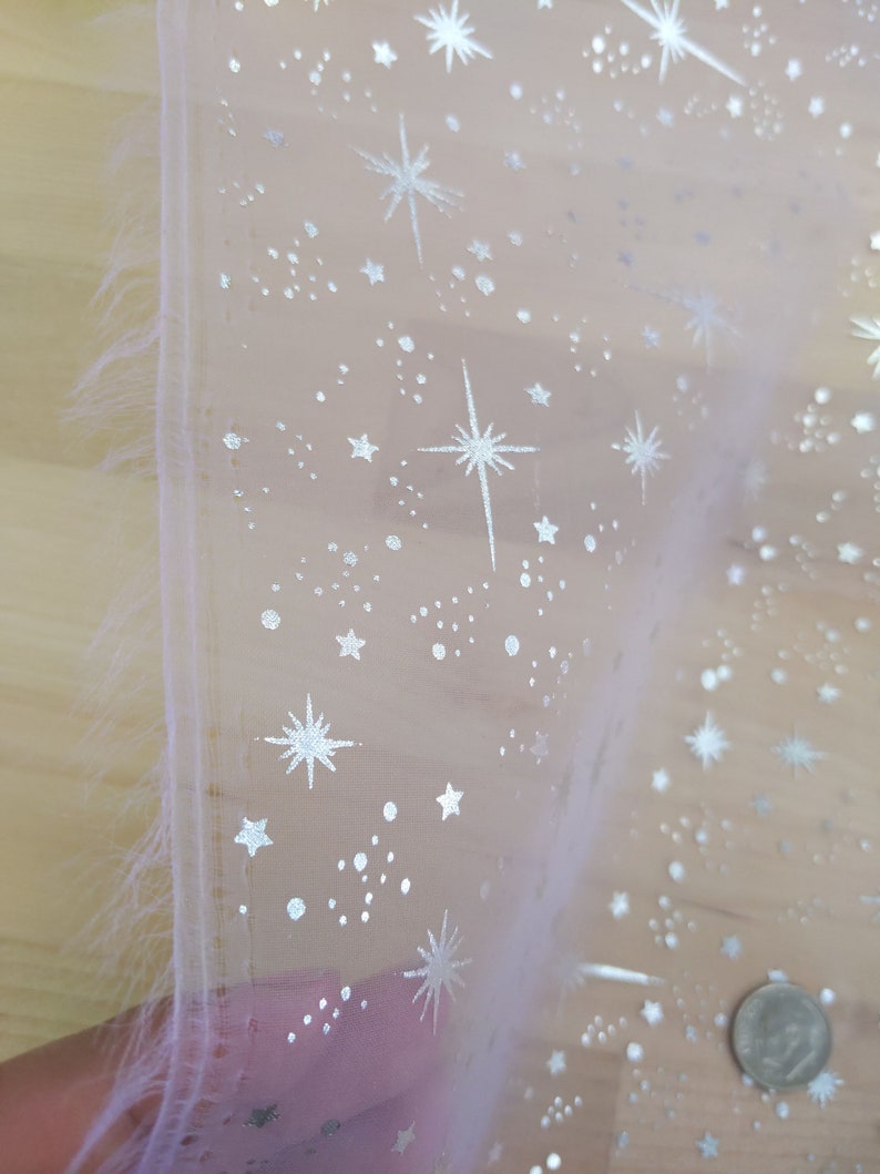 May include: A sheer white fabric with a silver glitter pattern of stars and dots. The fabric is folded in half and the edge is frayed.