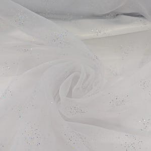 May include: White tulle fabric with scattered iridescent glitter. The fabric is draped and folded, creating soft shadows and textures. The glitter catches the light, adding a subtle sparkle. Suitable for crafting, bridal wear, or decorative projects.
