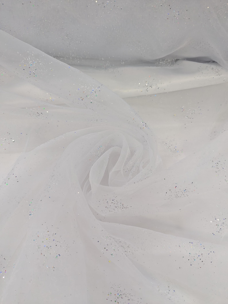 May include: White tulle fabric with scattered iridescent glitter. The fabric is draped and folded, creating soft shadows and textures. The glitter catches the light, adding a subtle sparkle. Suitable for crafting, bridal wear, or decorative projects.