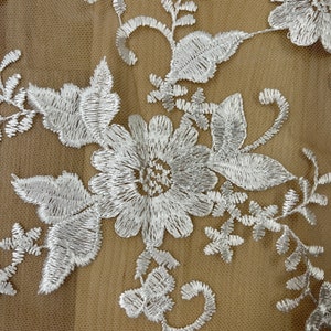 May include: Close-up of white floral embroidery on a beige mesh fabric. The embroidery features detailed flowers, leaves, and swirling vines. The delicate design is intricately stitched, creating a textured and elegant pattern.
