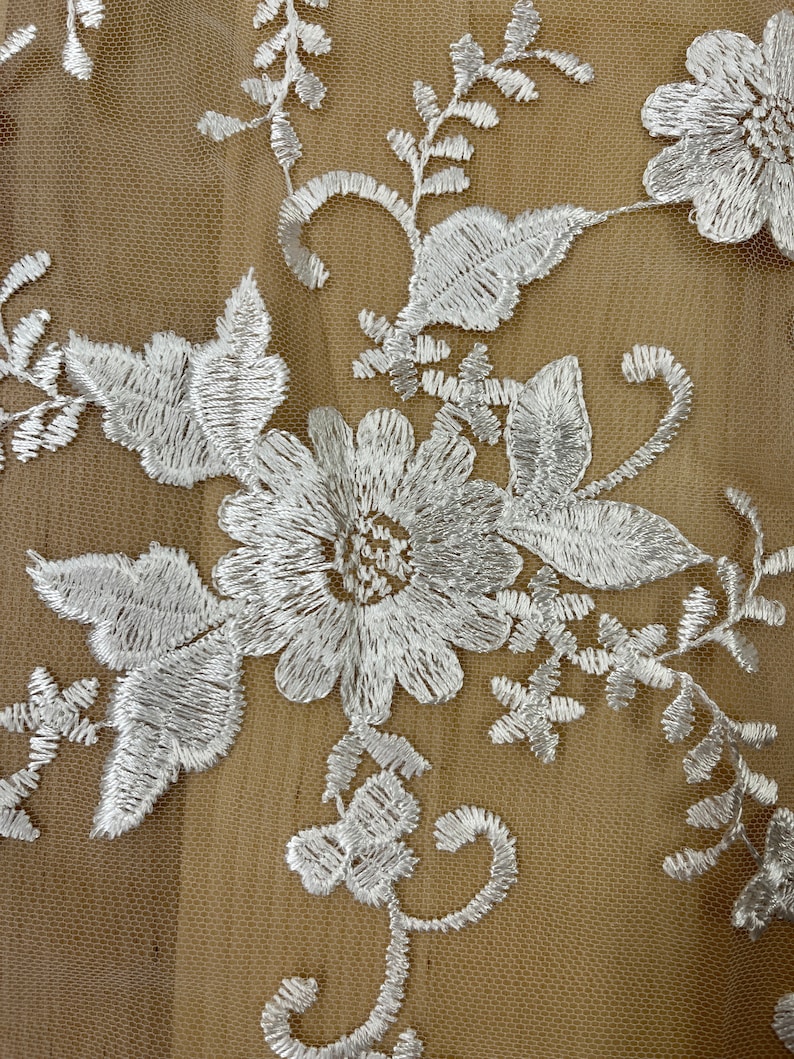 May include: Close-up of white floral embroidery on a beige mesh fabric. The embroidery features detailed flowers, leaves, and swirling vines. The delicate design is intricately stitched, creating a textured and elegant pattern.