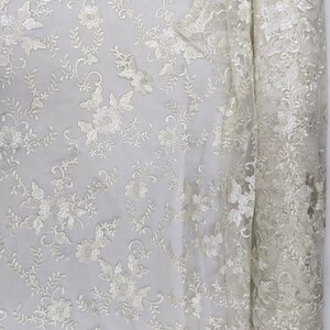 May include: Ivory-colored lace fabric with a floral pattern. The fabric is rolled up on the right side, revealing the intricate details of the embroidered flowers and leaves. The scalloped edge adds a delicate touch to the design.