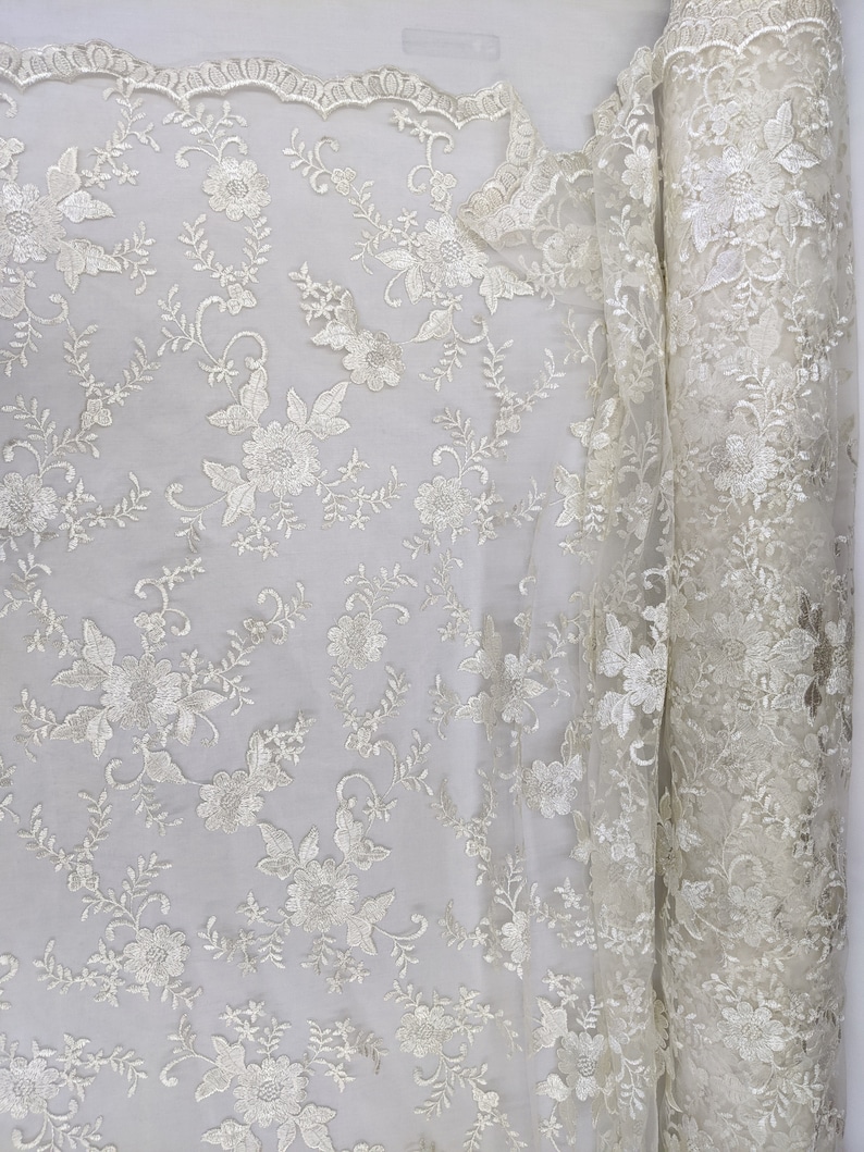 May include: Ivory-colored lace fabric with a floral pattern. The fabric is rolled up on the right side, revealing the intricate details of the embroidered flowers and leaves. The scalloped edge adds a delicate touch to the design.