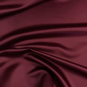 May include: A close-up shot of a deep burgundy fabric with a satin finish. The material is draped, creating soft folds and shadows that highlight the sheen. The fabric's rich color and smooth texture suggest it could be used for apparel or home decor.
