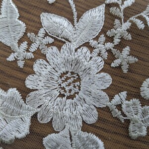 May include: Close-up of white floral lace fabric. The design features embroidered flowers, leaves, and delicate details. The lace is set against a brown background, highlighting the intricate patterns and textures.