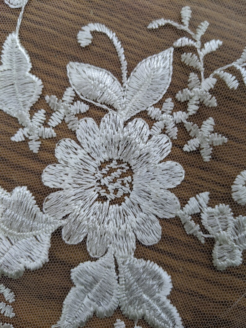 May include: Close-up of white floral lace fabric. The design features embroidered flowers, leaves, and delicate details. The lace is set against a brown background, highlighting the intricate patterns and textures.