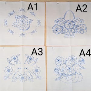 May include: Four white fabric squares with blue embroidered designs. A1 features a rose in a floral wreath. A2 shows a basket of roses. A3 depicts two birds with flowers. A4 displays a duck with roses. Each square has a red border.
