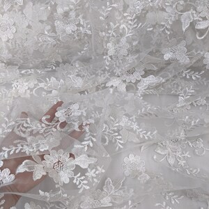 May include: Close-up of white floral lace fabric. The lace features detailed embroidered flowers and leaves, creating a delicate and textured pattern. The fabric is semi-sheer, allowing light to pass through, and is suitable for various craft and design projects.