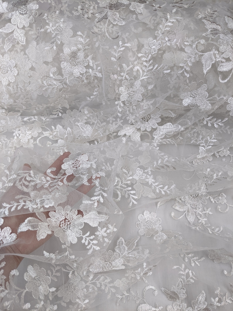 May include: Close-up of white floral lace fabric. The lace features detailed embroidered flowers and leaves, creating a delicate and textured pattern. The fabric is semi-sheer, allowing light to pass through, and is suitable for various craft and design projects.