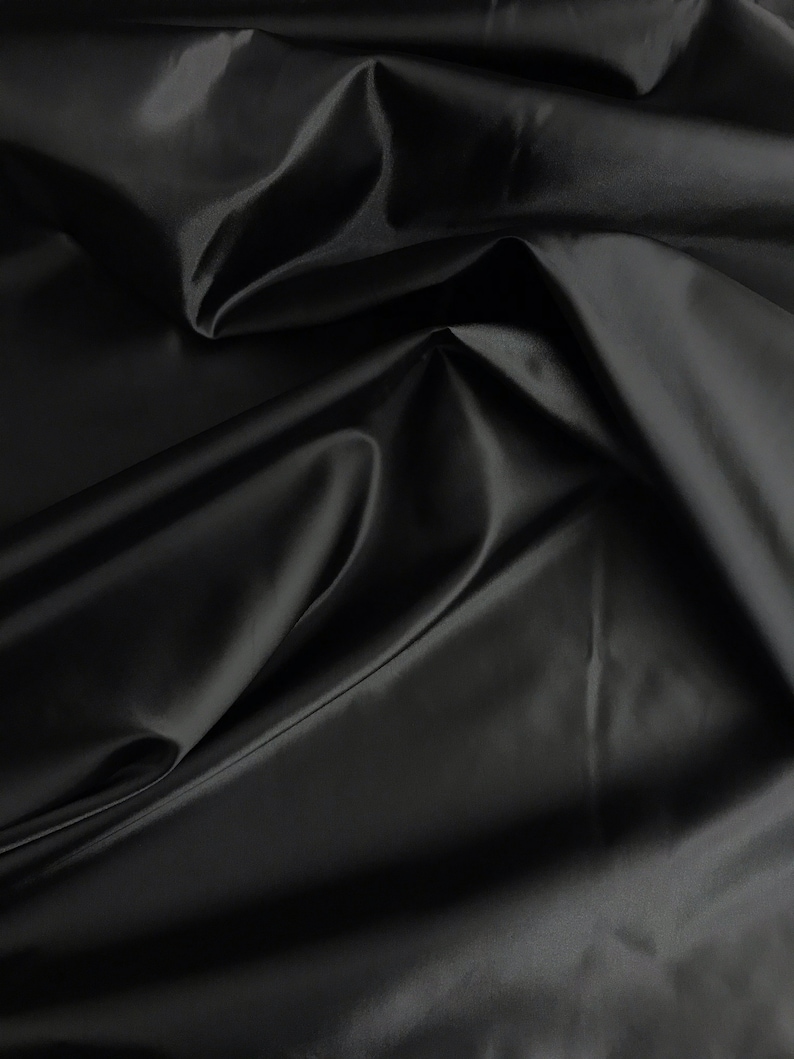 May include: Black satin fabric with a smooth, silky texture. The fabric is draped and folded, creating soft curves and folds.