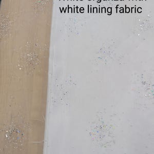 May include: White organza fabric with white lining, scattered with iridescent glitter. The text "White organza with white lining fabric" is visible on the fabric. This fabric is suitable for crafting and sewing projects.