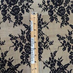 May include: Black floral lace fabric with a repeating pattern of roses and leaves. The fabric is sheer and delicate, with a soft, flowing texture. A ruler is included for scale, showing the fabric is 11 inches wide.