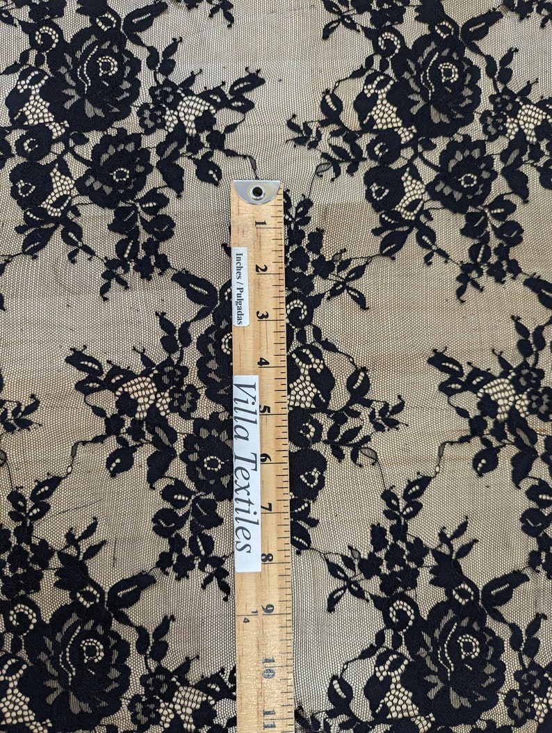 May include: Black floral lace fabric with a repeating pattern of roses and leaves. The fabric is sheer and delicate, with a soft, flowing texture. A ruler is included for scale, showing the fabric is 11 inches wide.
