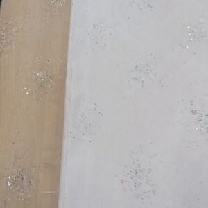 May include: White fabric with scattered iridescent glitter. The fabric is folded over, revealing a light brown surface. The glitter sparkles, creating a delicate, festive appearance.
