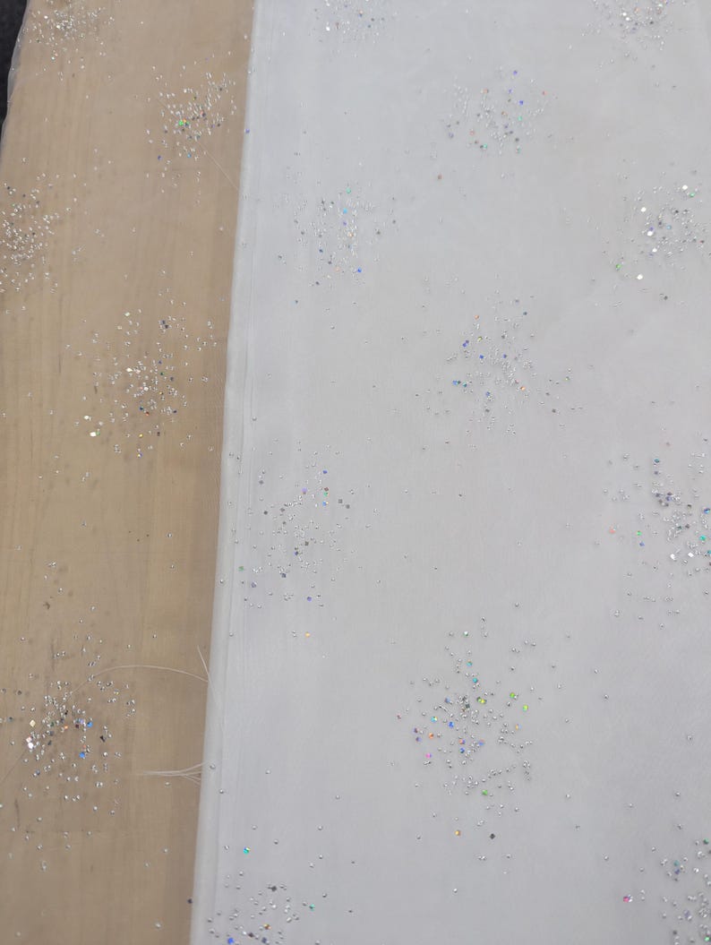 May include: White fabric with scattered iridescent glitter. The fabric is folded over, revealing a light brown surface. The glitter sparkles, creating a delicate, festive appearance.