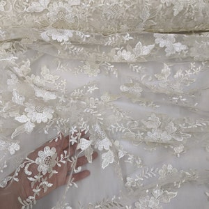 May include: Ivory-colored floral lace fabric. The delicate lace features embroidered flowers and leaves. The fabric is rolled up, showcasing the intricate details and texture. Ideal for bridal wear, dresses, or decorative projects.