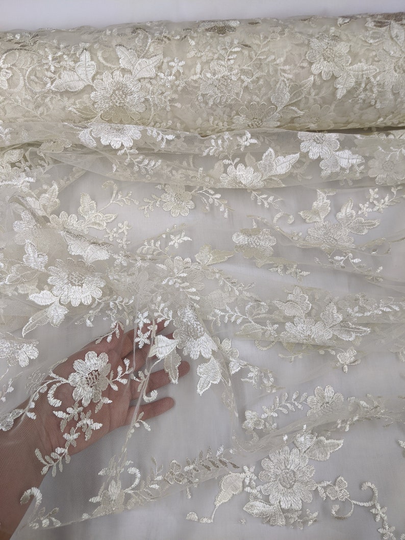 May include: Ivory-colored floral lace fabric. The delicate lace features embroidered flowers and leaves. The fabric is rolled up, showcasing the intricate details and texture. Ideal for bridal wear, dresses, or decorative projects.