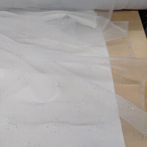 May include: White tulle fabric with scattered iridescent glitter. The fabric is draped and rolled, showing its delicate texture and potential for crafting. The glitter adds a subtle sparkle, enhancing its visual appeal. Ideal for bridal wear or decorative projects.