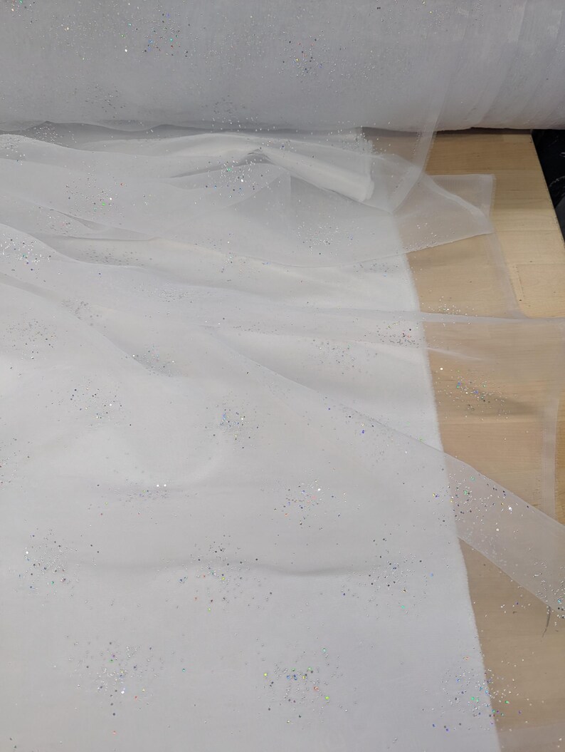 May include: White tulle fabric with scattered iridescent glitter. The fabric is draped and rolled, showing its delicate texture and potential for crafting. The glitter adds a subtle sparkle, enhancing its visual appeal. Ideal for bridal wear or decorative projects.