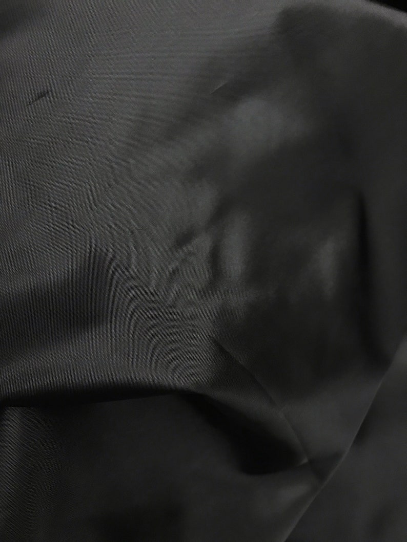 May include: Close-up of a black fabric with a smooth, slightly shiny texture. The fabric is wrinkled and folded, showing its drape and flexibility.