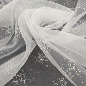 May include: White, translucent fabric sprinkled with iridescent glitter. The material is arranged in flowing folds, producing gentle curves and shadows. The glitter reflects light, creating a shimmering effect. Suitable for crafts, decorations, or clothing.