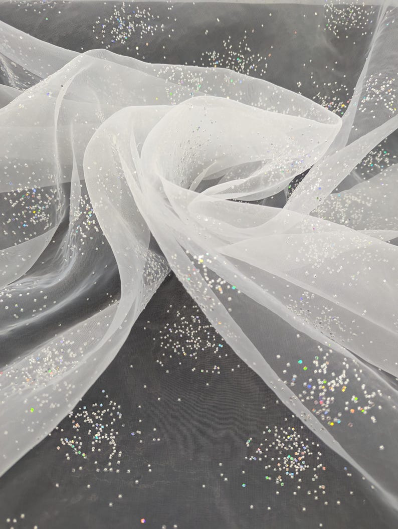 May include: White, translucent fabric sprinkled with iridescent glitter. The material is arranged in flowing folds, producing gentle curves and shadows. The glitter reflects light, creating a shimmering effect. Suitable for crafts, decorations, or clothing.