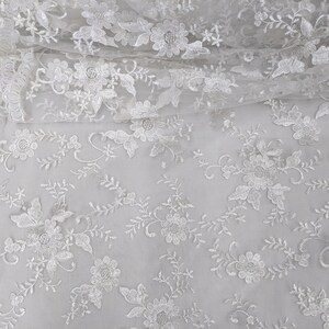 May include: White floral lace fabric with detailed embroidered flowers and leaves. The delicate lace features a scalloped edge and is suitable for bridal wear, dresses, or craft projects. The fabric is lightweight and semi-sheer, perfect for overlays.
