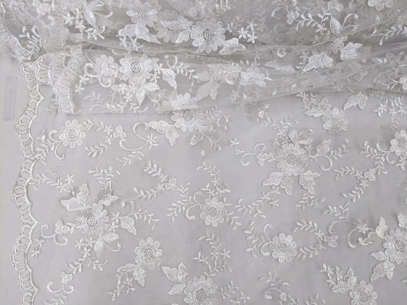 May include: White floral lace fabric with detailed embroidered flowers and leaves. The delicate lace features a scalloped edge and is suitable for bridal wear, dresses, or craft projects. The fabric is lightweight and semi-sheer, perfect for overlays.