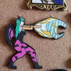 May include: A black, pink, and green enamel pin of a female character with wings, wearing a black dress and pink leggings with bat designs. She is holding a yellow and blue striped object in her hand.