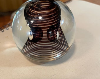 May include: A clear glass paperweight with a swirling, dark brown design inside. The design is a series of concentric circles that create a 3D effect.