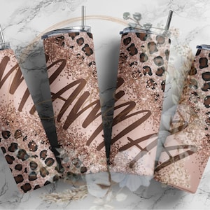 Pode incluir: Quatro copos de aço inoxidável com tampas e canudos. Os copos são decorados com um fundo rosa, glitter dourado e estampa de leopardo. A palavra "Mama" está escrita em letras marrons em dois dos copos.