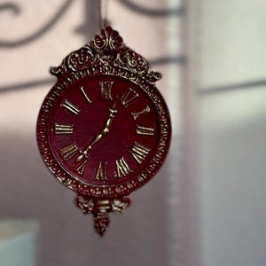 May include: A circular wall clock featuring a deep red face with raised gold Roman numerals and matching gold hands. The clock is encased in an ornate, decorative gold-trimmed frame with intricate scrollwork details at the top and bottom.