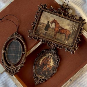 May include: Three decorative hanging ornaments with ornate, dark bronze-colored frames. One rectangular frame features a person standing with a brown horse, one oval frame contains a brown horse portrait, and one oval frame displays a dark plaid pattern.